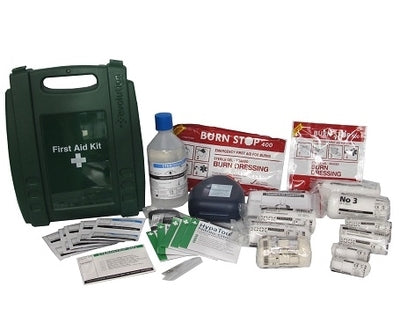 A green first aid box with various supplies including adhesive plasters, sterile eye pads, disposable triangular bandages, safety pins, wound dressings and crepe bandages, laid out on a white background.